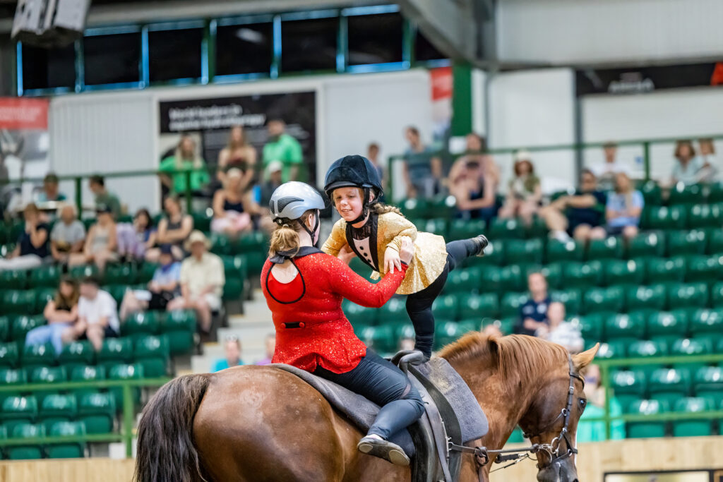 Lizzie and vaulter Eden are on a horse with Lizzie supporting Eden as she stands and stretches a leg behind her