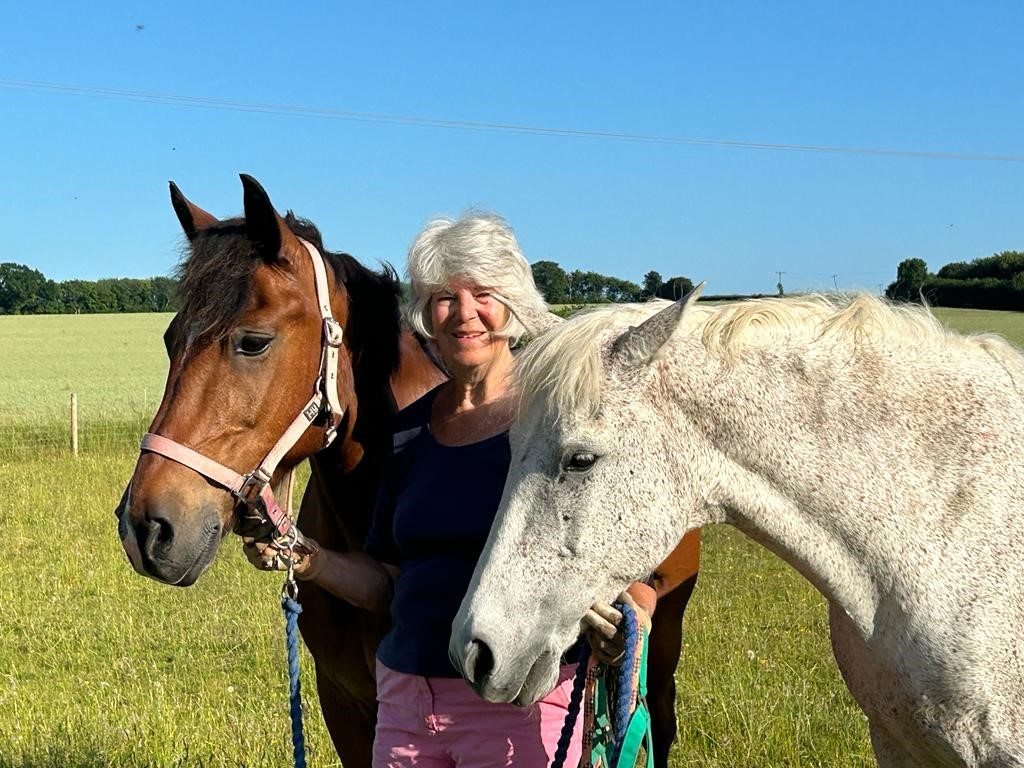 Catherine stands in a field with two horses either side of her. It is a bright sunny, summer day with blue skies. The horse on her left is a bay horse and on the right is a grey horse.