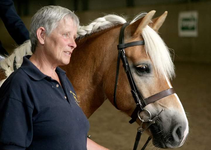 Jane Barker stands next to a palomino horse n a side profile, looking off to the right. Jane is holding the horse's reins out of the camera's view. The horse has a plaited mane.