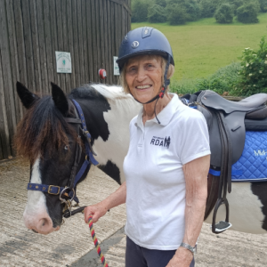 Ruby stands with a saddled piebald pony, she's wearing a white polo and riding hat smiling to the camera.