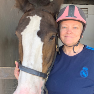 Participant of the Year Sheila stands next to a horse in a stable. Sheila is touching its pink nose and smiling at the camera. Sheila is wearing a pink riding hat and the horse is a bay horse with a white blaze.
