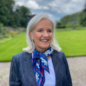 Barbara Manson stands in a blue suit jacket, blue top and blue patterned silk scarf against a pristine lawn and garden.