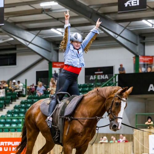 Lizzie Bennett on horse Puro standing up in the stirrups and with her hands raised in the air. She is wearing black riding trousers and a tassled blue jacket.