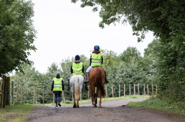 Two riders and a volunteer walk down a track surrounded by woodland.