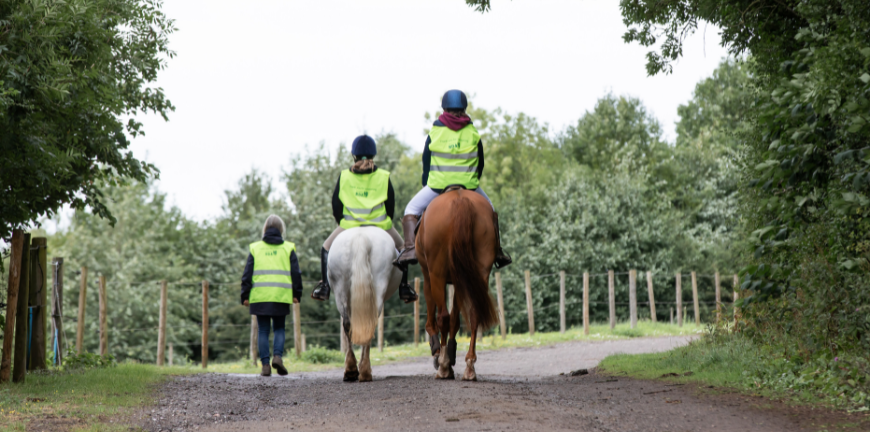 Two people riding horses, one on a grey pony and one on a chestnut horse, with someone walking alongside them along a country road. They are all in high vis jackets.