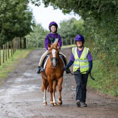 Participant is riding a chestnut pony and is escorted by a volunteer in a high vis. Both are walking down a track.