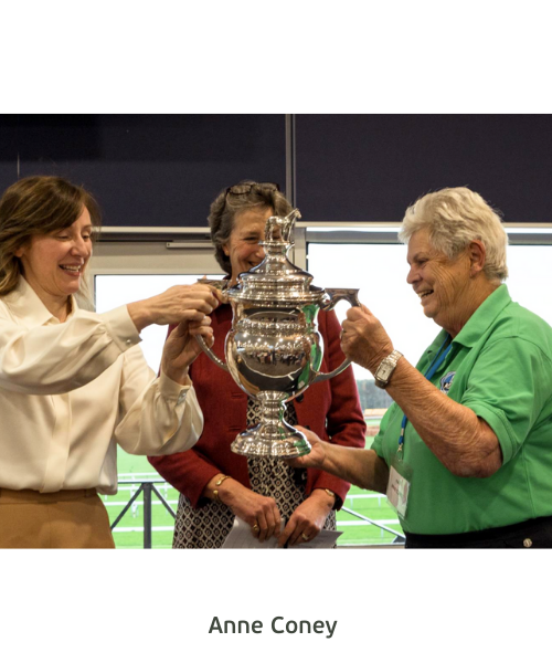 A woman in a green polo shirt receives a large silver trophy. Two women smile as she receives her award.