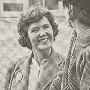 Sheila smiles at a rider. She has curly hair and a blazer with a brooch.