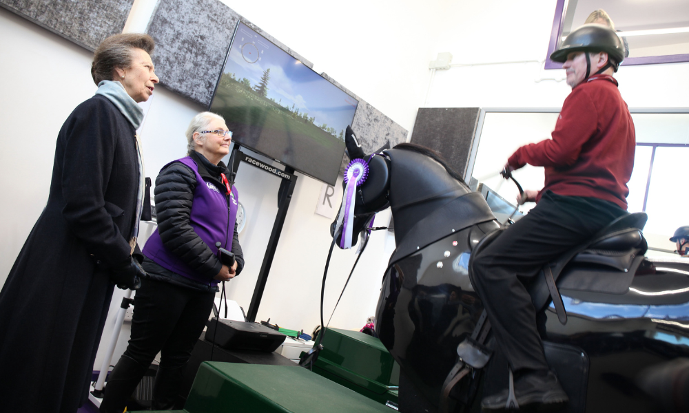 Rider Tim sits on top of a black mechanical horse while coach Many and the Princess Royal watch.