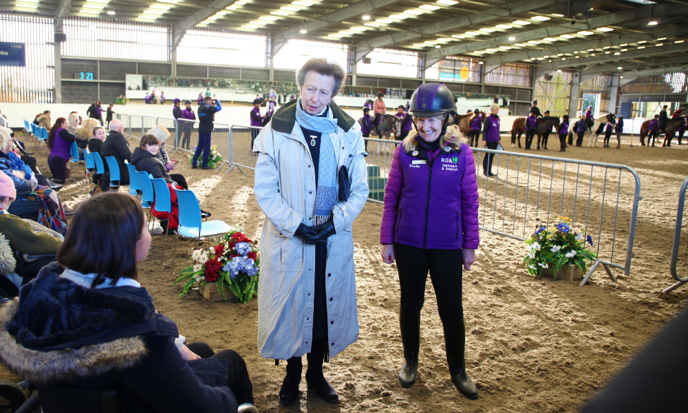 The Princess Royal stands next to Sheila Saner in a long white coat. They are in an indoor equestrian school talking to riders and familys of Nantwich & District RDA.
