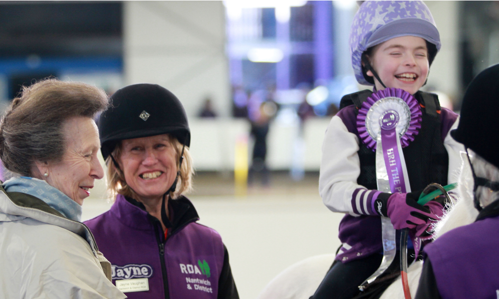 Rider Esme sits on top of a pony laughing with a large purple rosette. Princess Anne and Jayne Vaughan chat.
