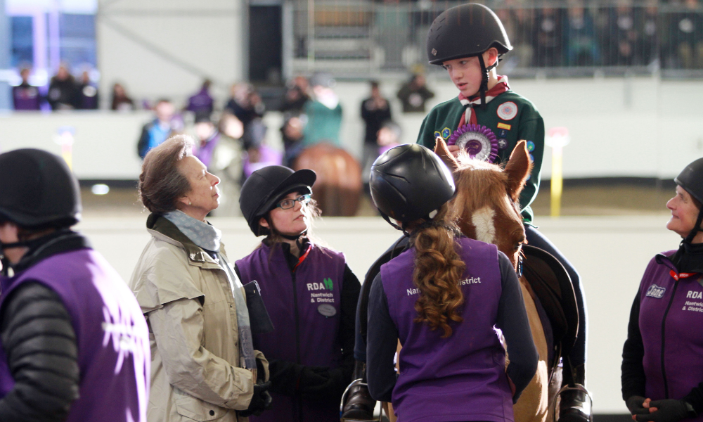Three volunteers in purple gilets and Princess Anne stand talking to rider Theo, as he sits on top of a horse.