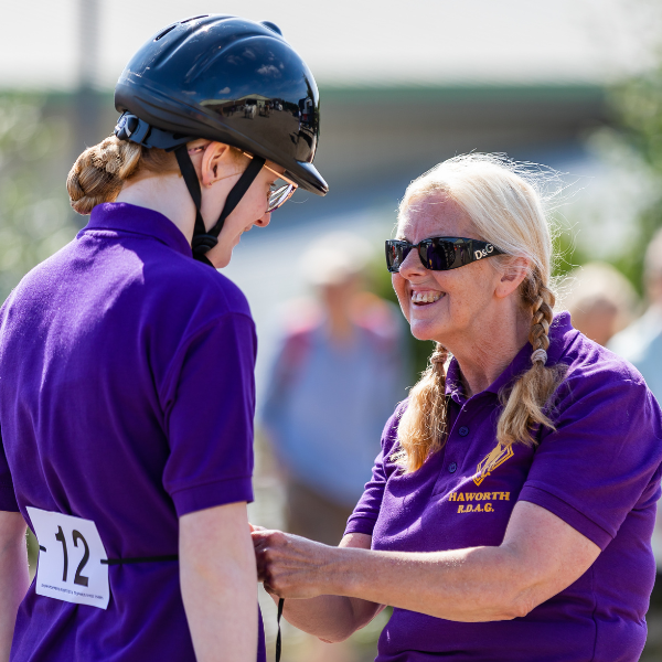 A volunteer and RDA rider chat to each other. The volunteer is tying a competition number around the riders waist,