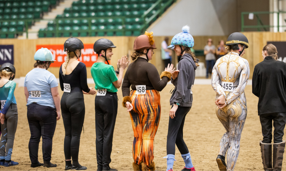 Eight vaulting competitors stand in a line in their competition outfits with a competitor number tied around their waist.