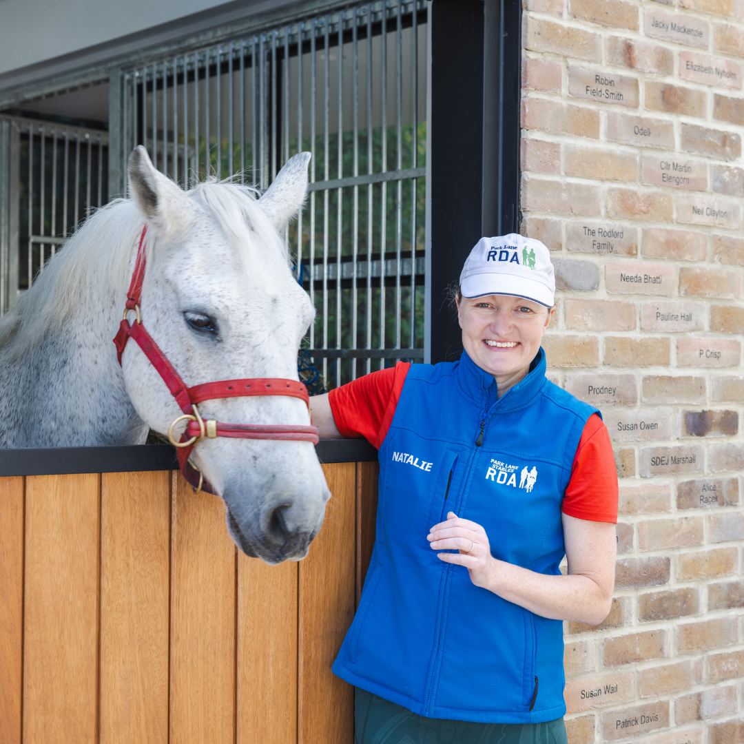 A women stood next to a grey horse wearing a red t-shirt, blue gilet and white hat, smiling towards the camera