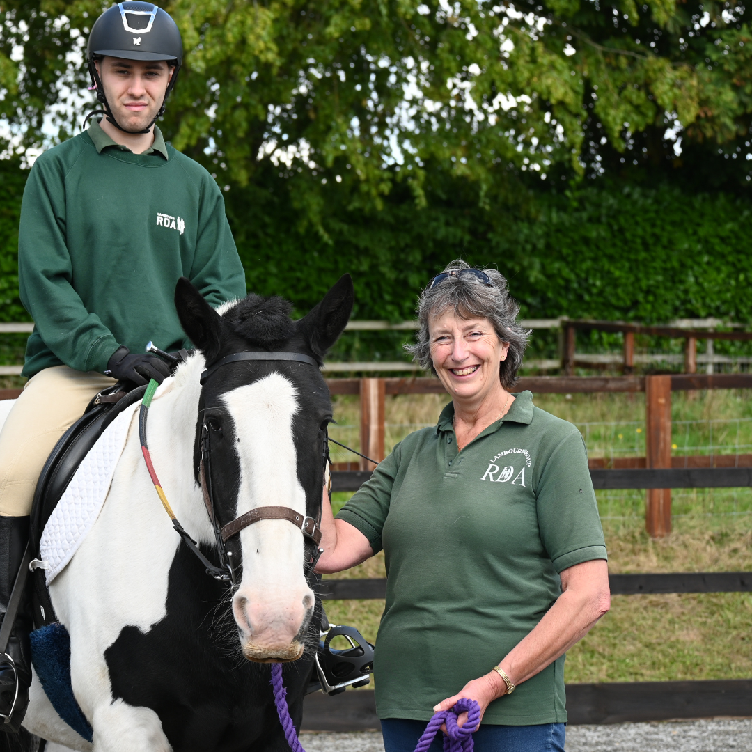 A man sat on a piebald horse with a women wearing a green polo stood next to him.