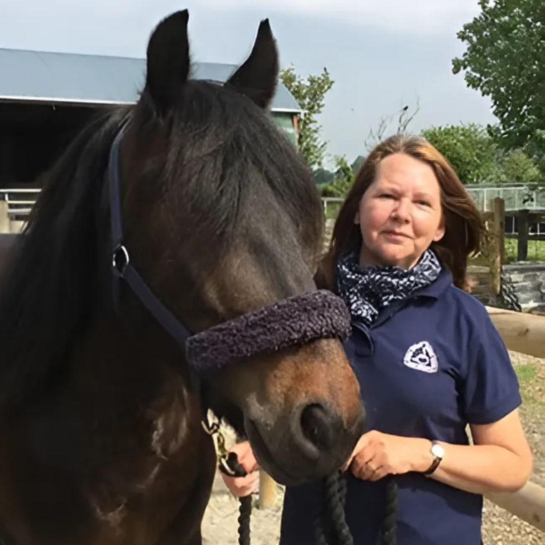 A woman wearing a blue polo shirt stood next a dark bay horse, smiling towards the camera