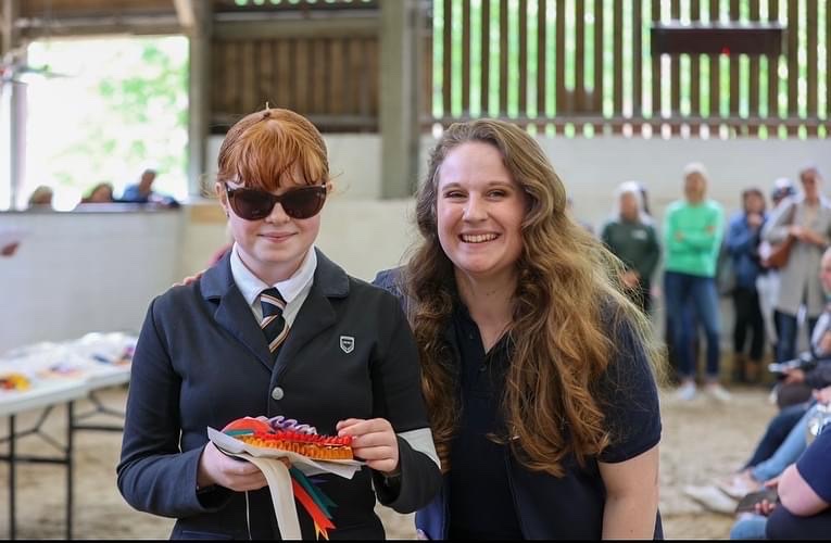 A visually impaired girl, wearing sunglasses, holding a rosette wile stood next to a lady wearing a dark navy shirt.