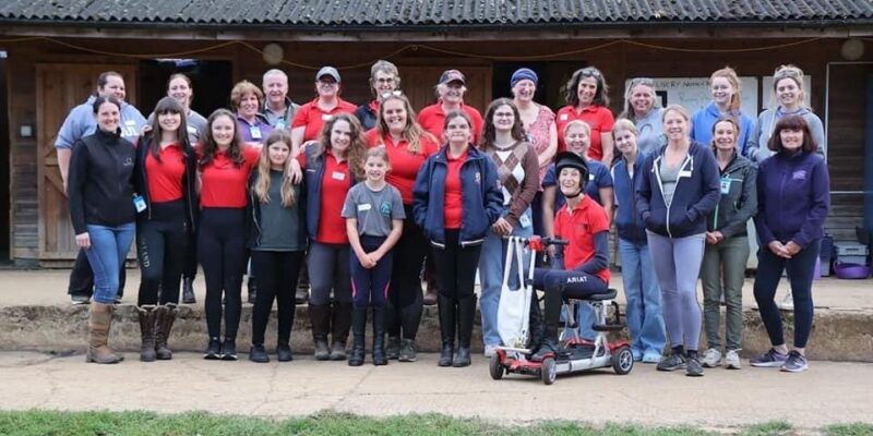 Aim RDA group stood together in front of the stables smiling for a group photo.