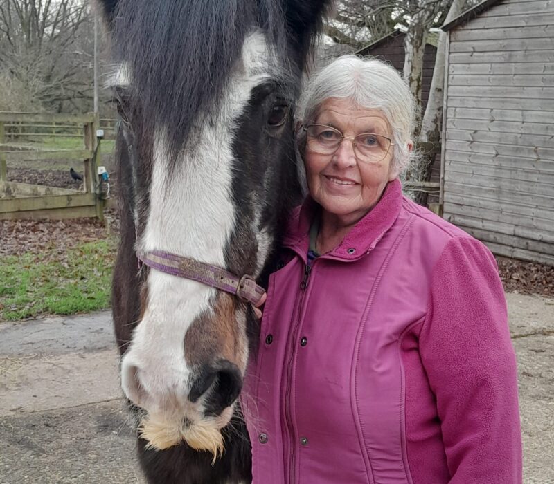 A older woman wearing a pink fleece, stood next to a horse, smiling at the camera.