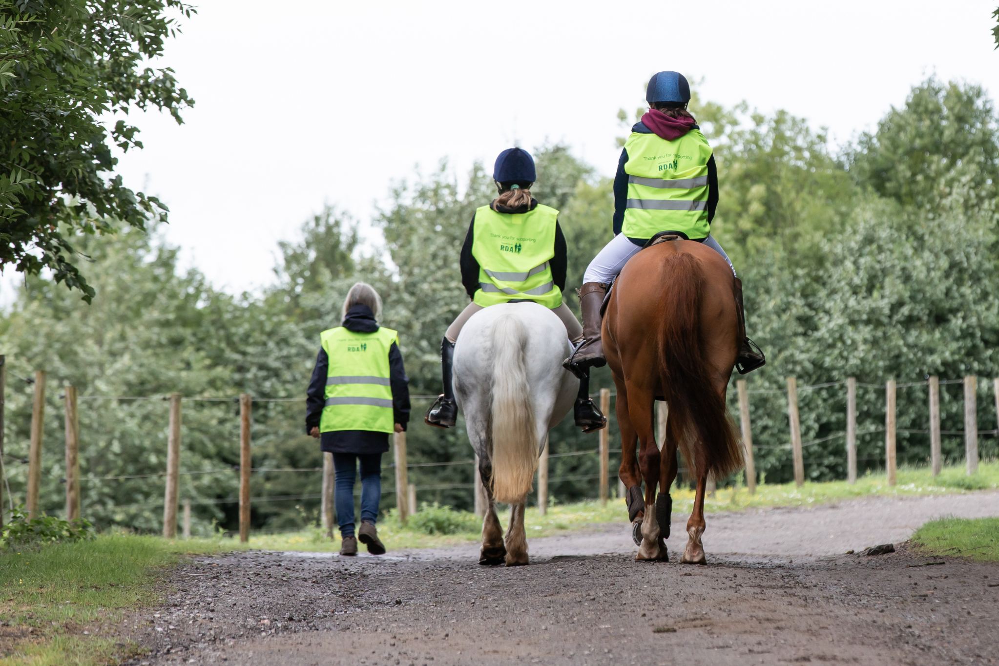 2 riders on horses and a coach walking along side them down a path