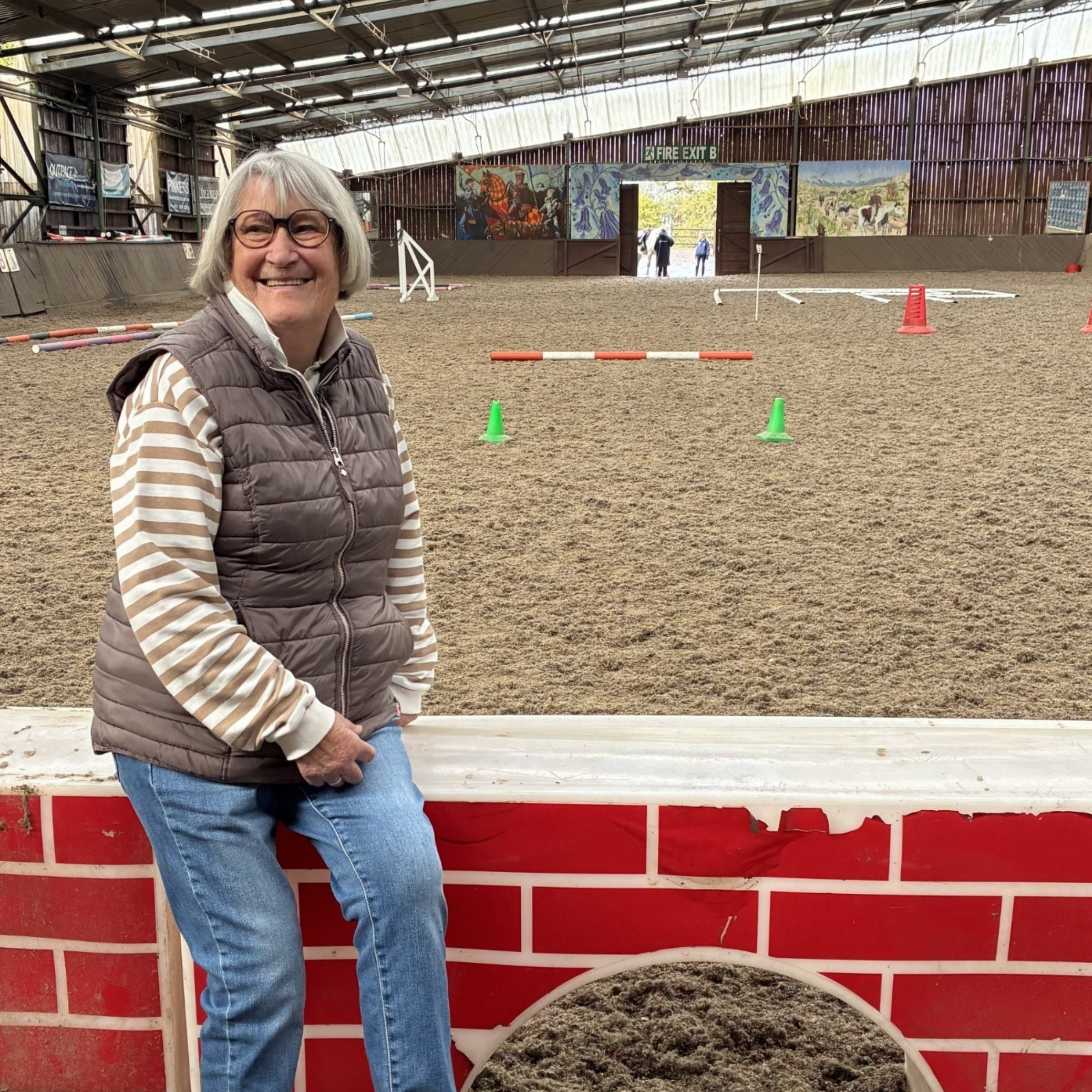 Sue Mack, sat on a fake brick wall, inside an indoor arena, smiling past the camera.