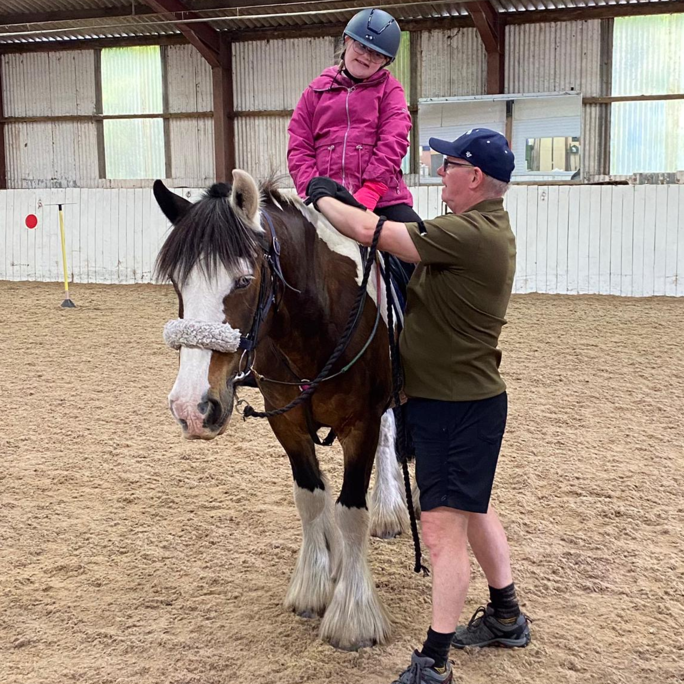 A man helping a young rider hold onto to reins. They are in a indoor arena