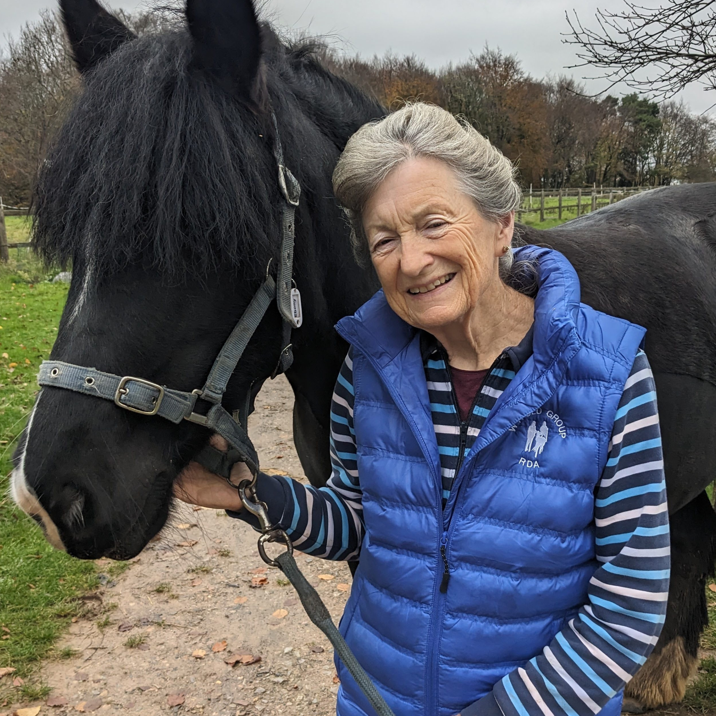 An older lady, in blue coat holding a black horse smiling at the camera.