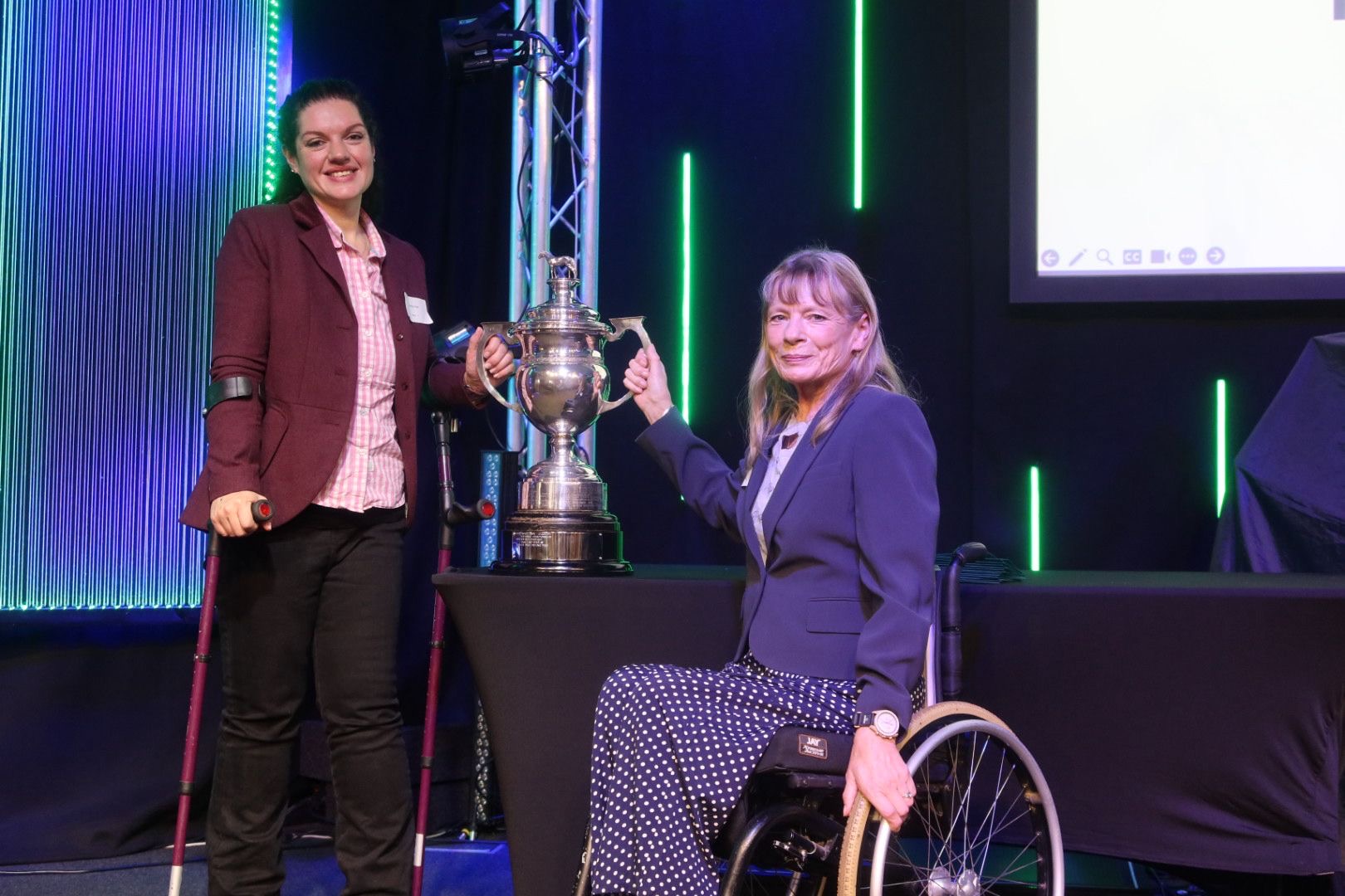 Two members of The British Para Driving Team collecting the Birt Spooner Cup on stage at the RDA AGM. They are holding the cup and smiling into the camera.